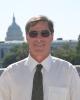 A man wearing a white shirt and dark tie stands smiling. The Capitol building is visible behind him.