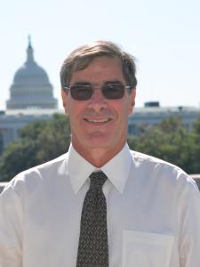 A man wearing a white shirt and dark tie stands smiling. The Capitol building is visible behind him.